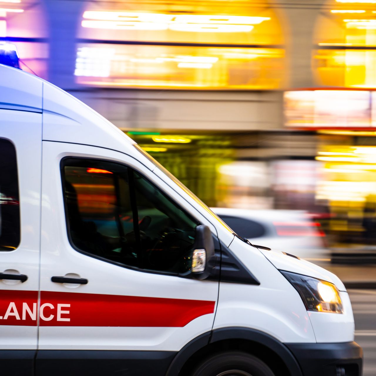 Ambulance blanche à bande rouge, gyrophare bleu allumé, filant à toute allure dans une rue floue.