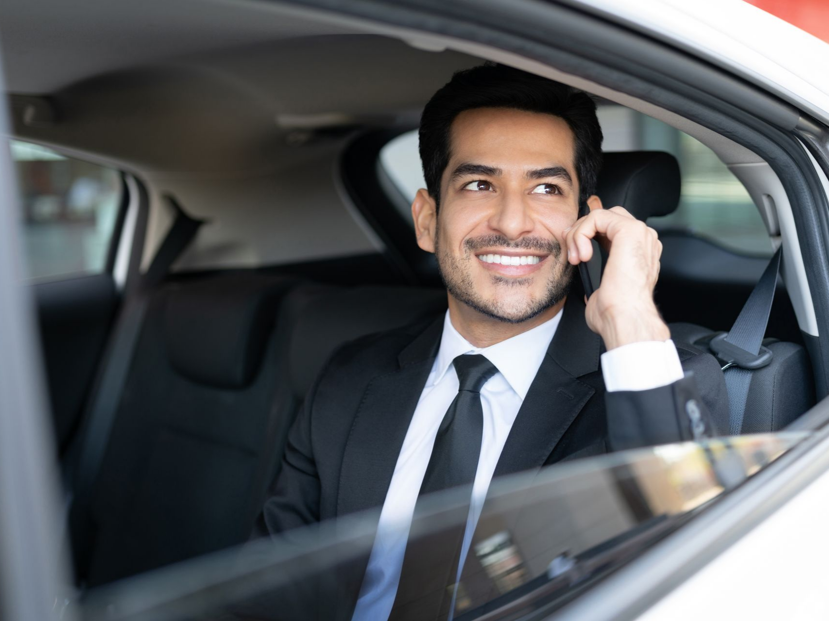 Un homme en costume sourit, parlant au téléphone sur la banquette arrière d'une voiture.