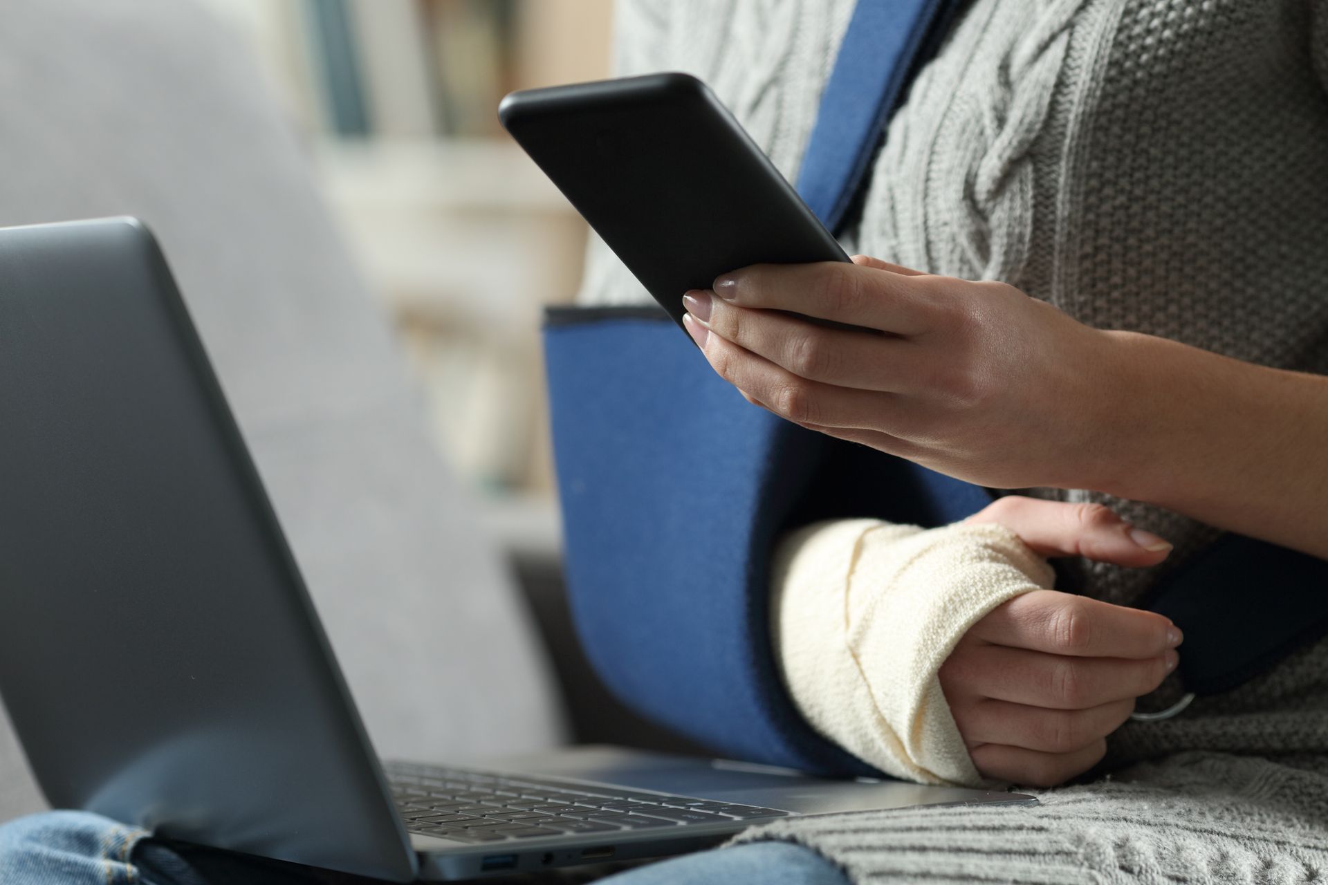Femme avec le bras en écharpe et le poignet bandé, utilisant un téléphone et un ordinateur portable.