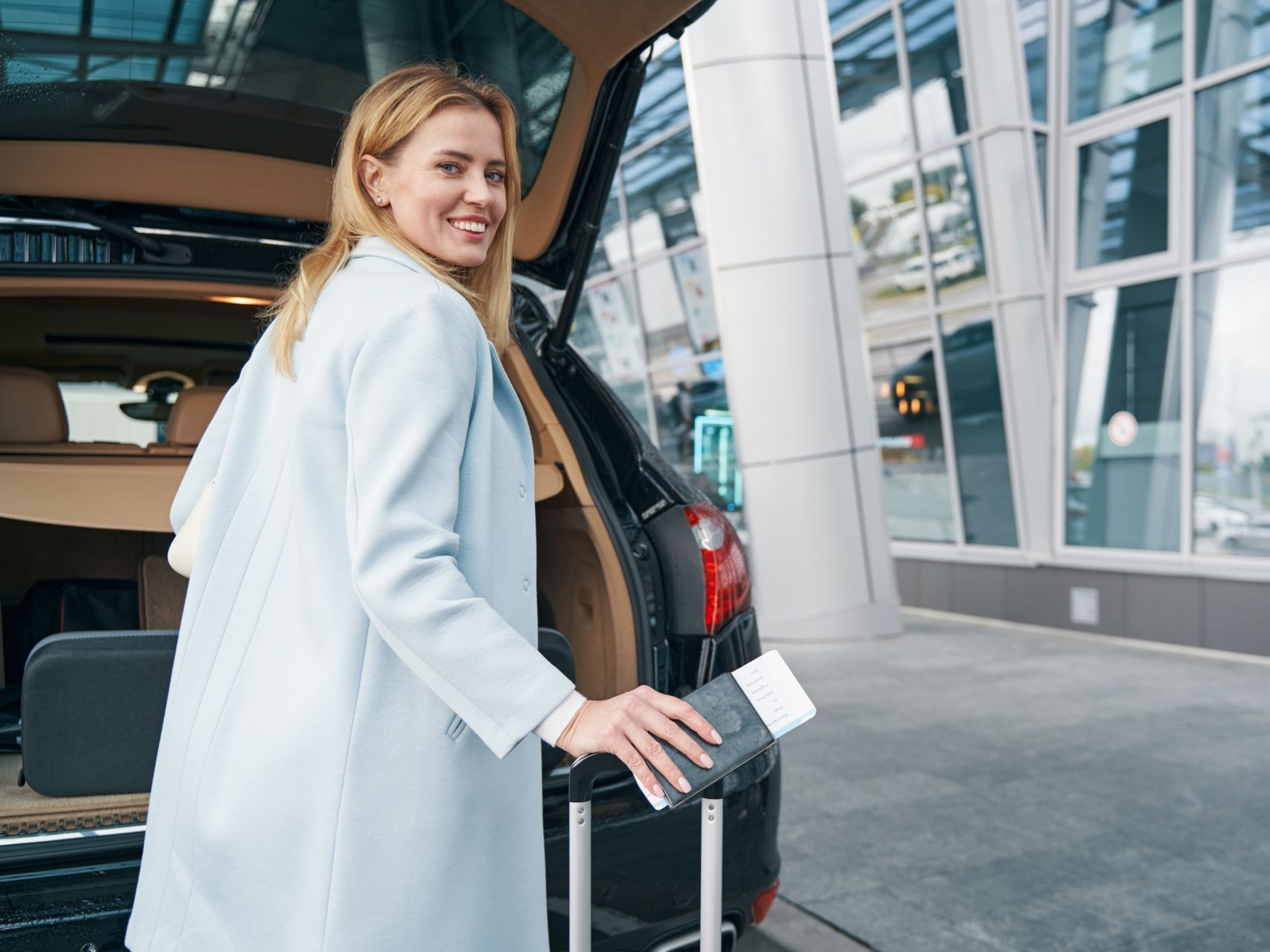 Une femme range ses bagages dans le coffre d'une voiture près d'un aéroport, tenant son passeport et souriant.