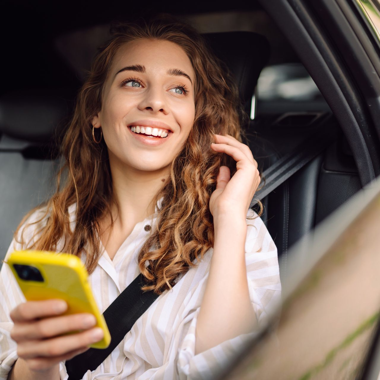 Une femme dans une voiture sourit, tenant un téléphone jaune, le regard tourné vers le ciel.
