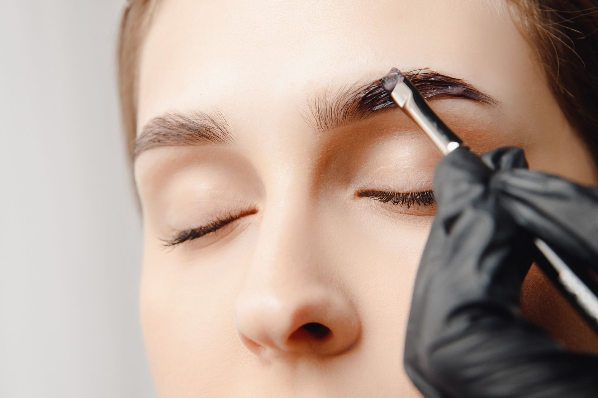 Mujer con los ojos cerrados, con una ceja teñida de oscuro con un pincel y una mano enguantada.