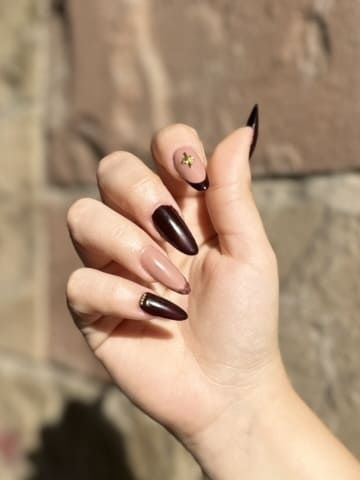 Mano con uñas largas en forma de almendra; algunas son de color burdeos, nude y adornadas con detalles dorados.