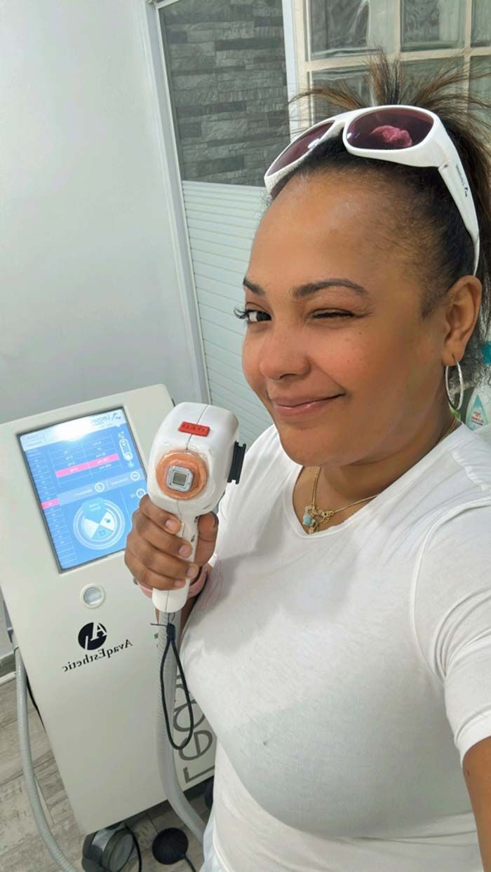 Mujer guiña un ojo, sostiene un dispositivo de depilación láser, una camiseta blanca y gafas de sol en la cabeza, en la clínica.