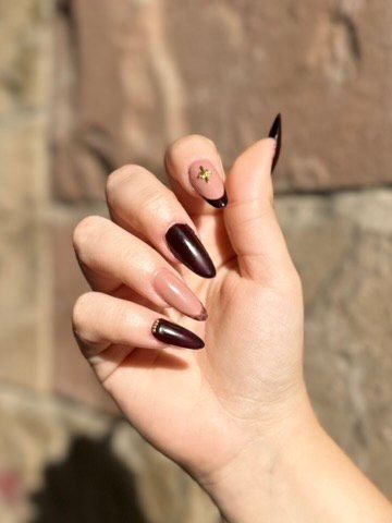 Mano con uñas almendradas, algunas color piel con puntas negras, otras color burdeos. Una uña tiene una cruz dorada.