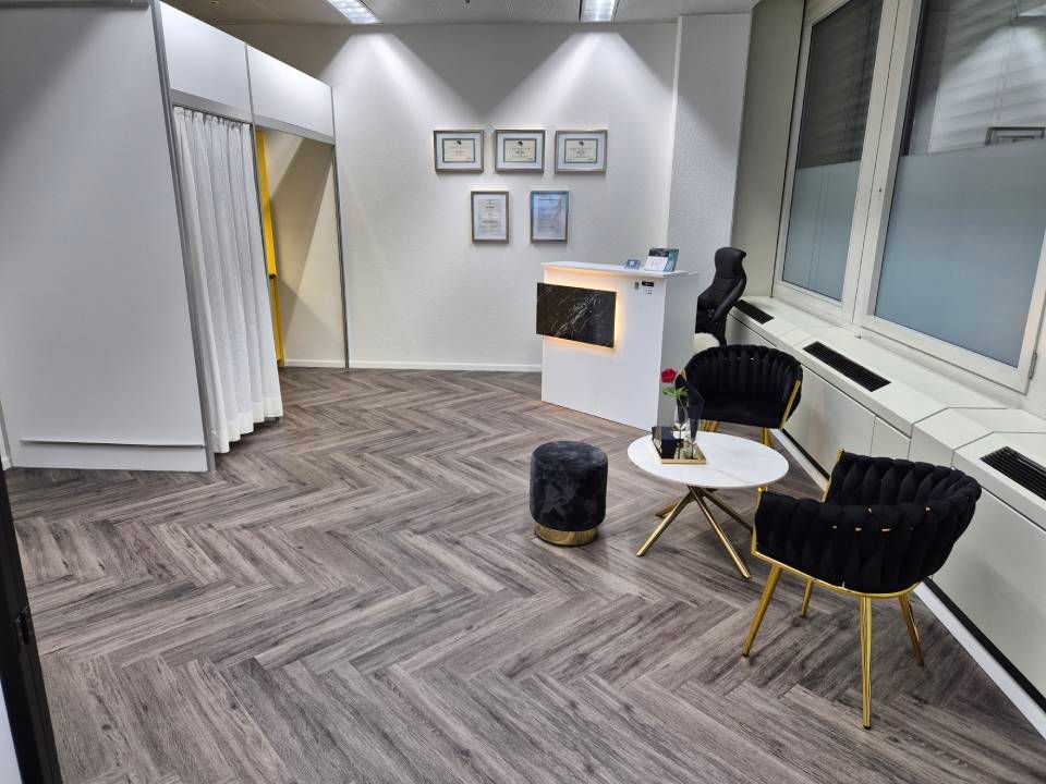 Ein Empfangsbereich mit weissen Wänden, Holzbodenoptik, einem Empfangstresen und einer Sitzecke mit zwei schwarzen Stühlen.