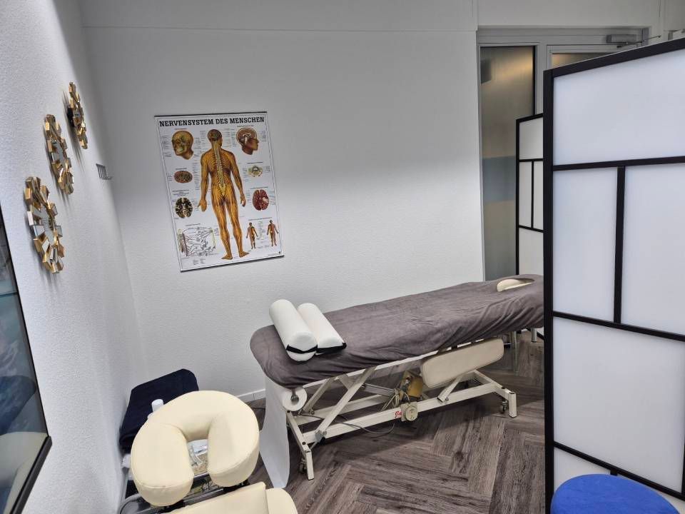 Ein klinischer Behandlungsraum mit Massageliege, Kissen und einer Anatomietafel an der Wand.