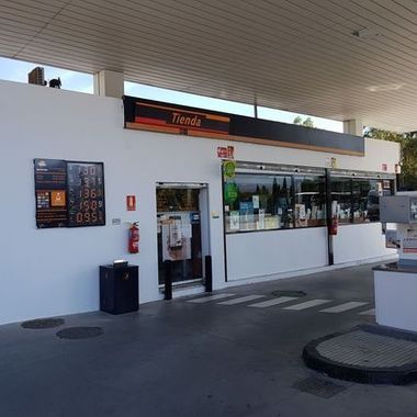 Exterior de una tienda de conveniencia de una gasolinera, con fachada blanca, ventanas con bebidas expuestas y zona de surtidores cubierta