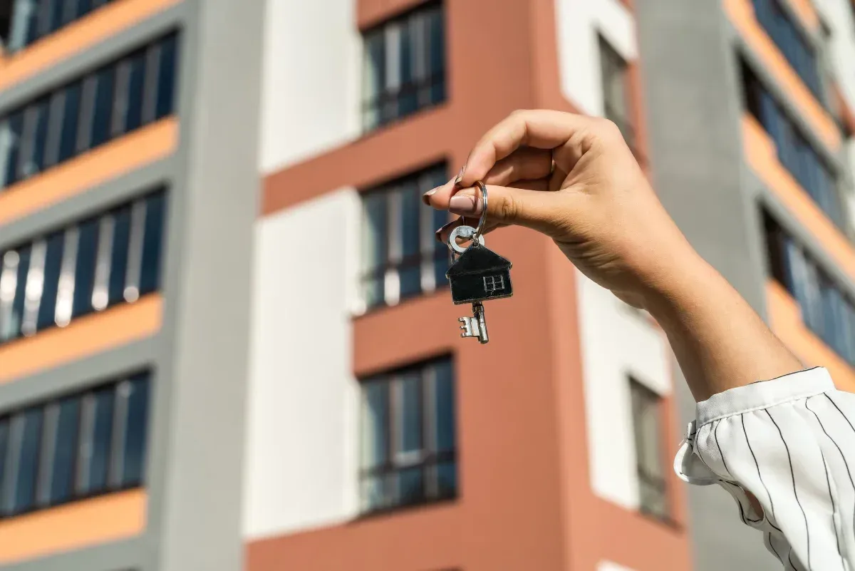 Mano sosteniendo llaves con un llavero en forma de casa frente a un edificio de apartamentos.