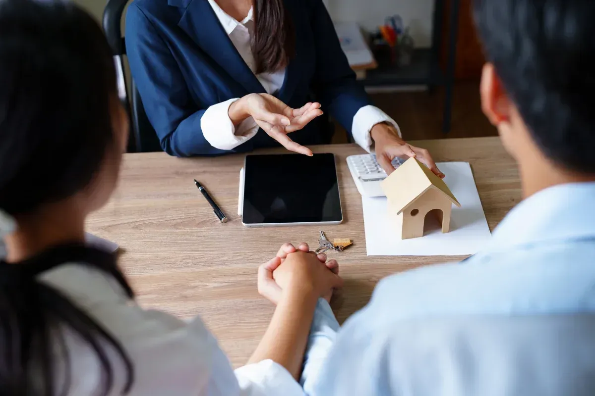 Una pareja y un agente inmobiliario discuten documentos en una mesa junto a una maqueta de casa.