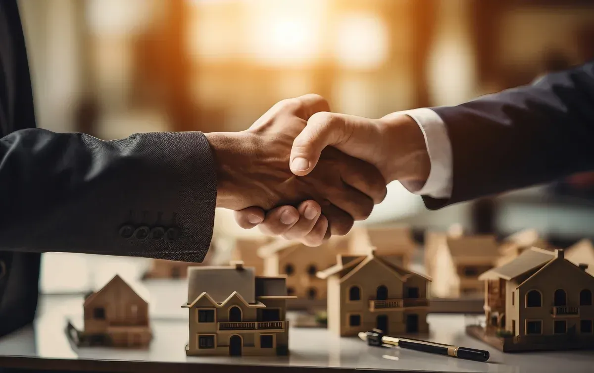 Dos personas con traje se dan la mano junto a maquetas de casas en miniatura.