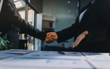 Dos personas con traje se dan la mano sobre una mesa con papeles.