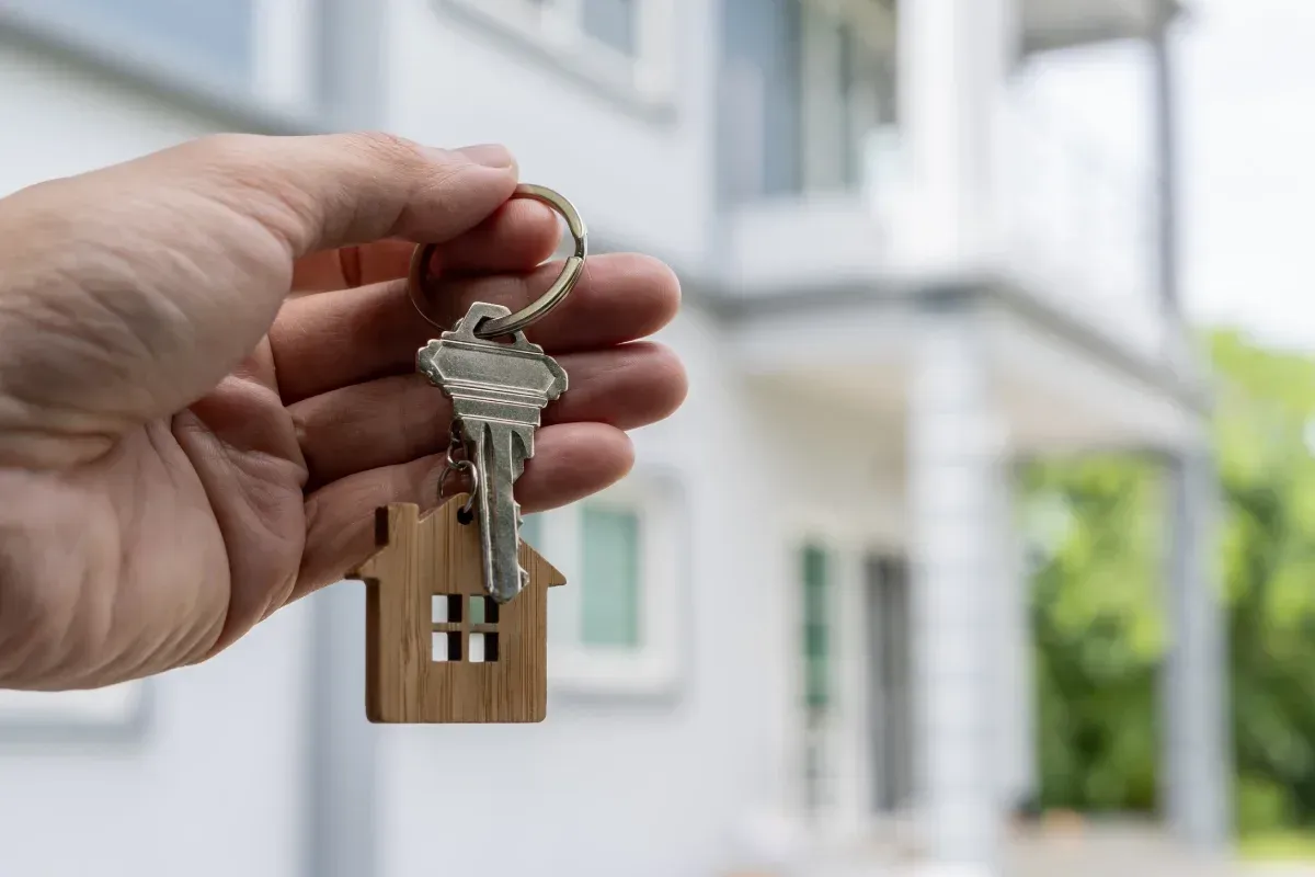 Mano sosteniendo las llaves de una casa frente a una vivienda moderna.