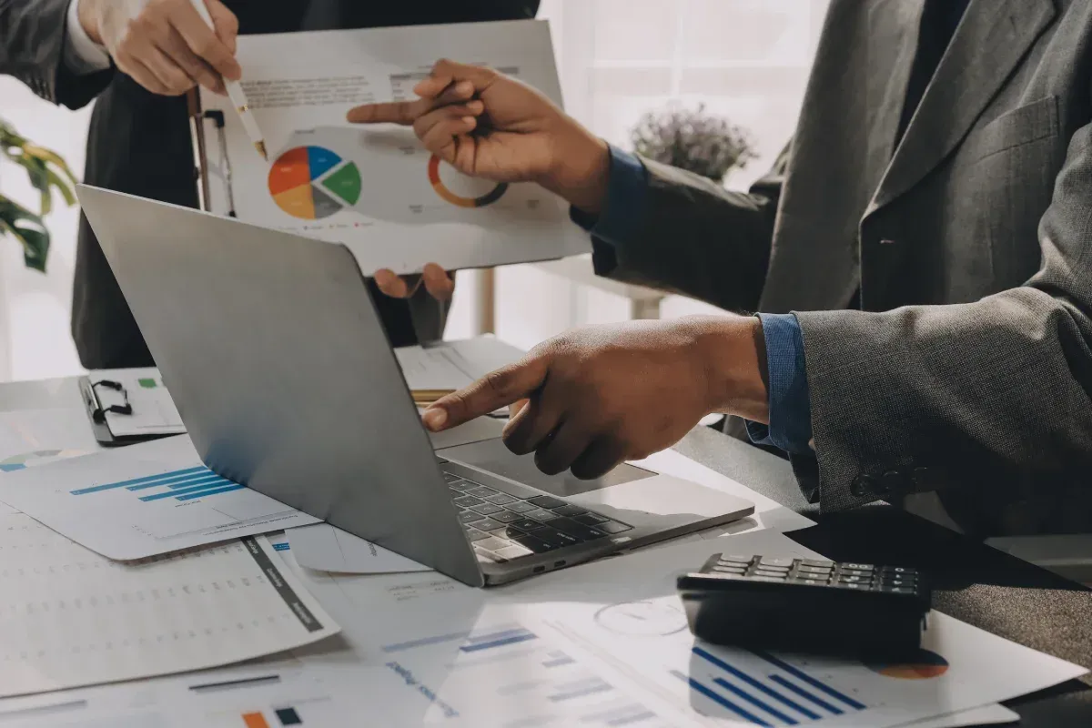 Dos personas vestidas de traje analizan datos financieros en un ordenador portátil y documentos con un gráfico circular.