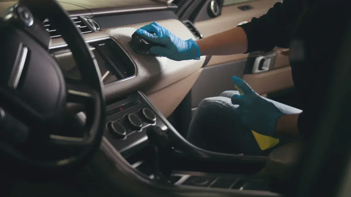 Persona con guantes azules limpiando el salpicadero beige de un coche. Se ven el volante y otros detalles del interior.