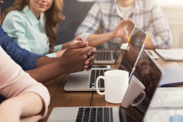 Personas sentadas a una mesa con computadoras portátiles, tomadas de la mano, en una reunión de trabajo colaborativa.