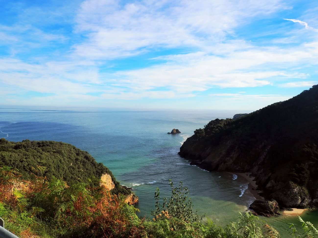Vista de una bahía turquesa con acantilados y playa bajo un cielo azul parcialmente nublado.