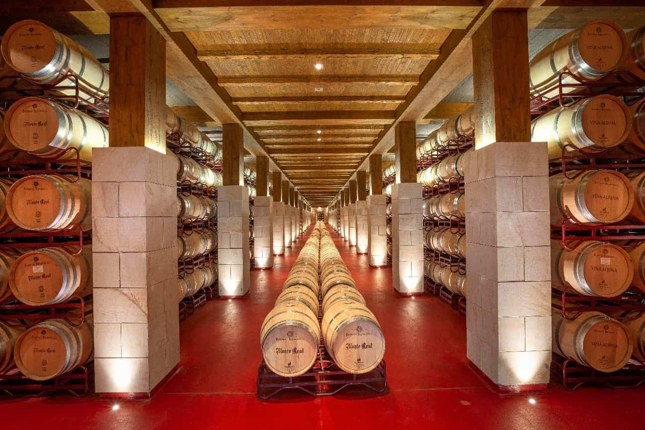 Los barriles de vino se alinean en una larga bodega con suelo rojo, columnas de piedra y techos de madera.