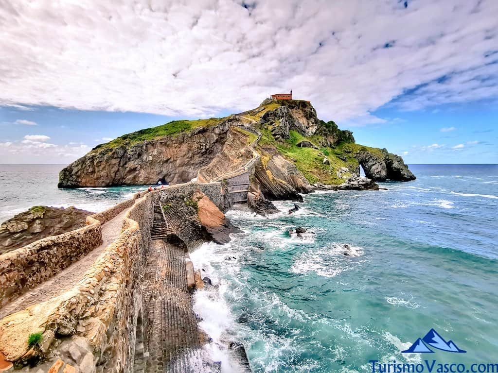 Isla con una pequeña capilla en la cima, conectada por un puente de piedra sobre aguas turquesas. Cielo nublado.