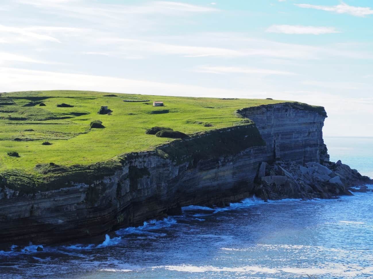 Un acantilado verde y herboso se encuentra con el océano azul bajo un cielo parcialmente nublado.
