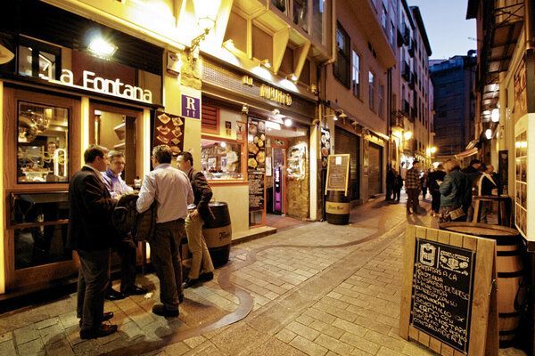 Calle con restaurantes y gente socializando al aire libre. Iluminada con luces cálidas y con un camino de piedra.