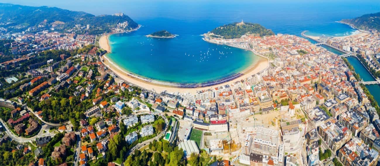 Vista aérea de San Sebastián, España, con una playa en forma de media luna, edificios y agua azul clara.