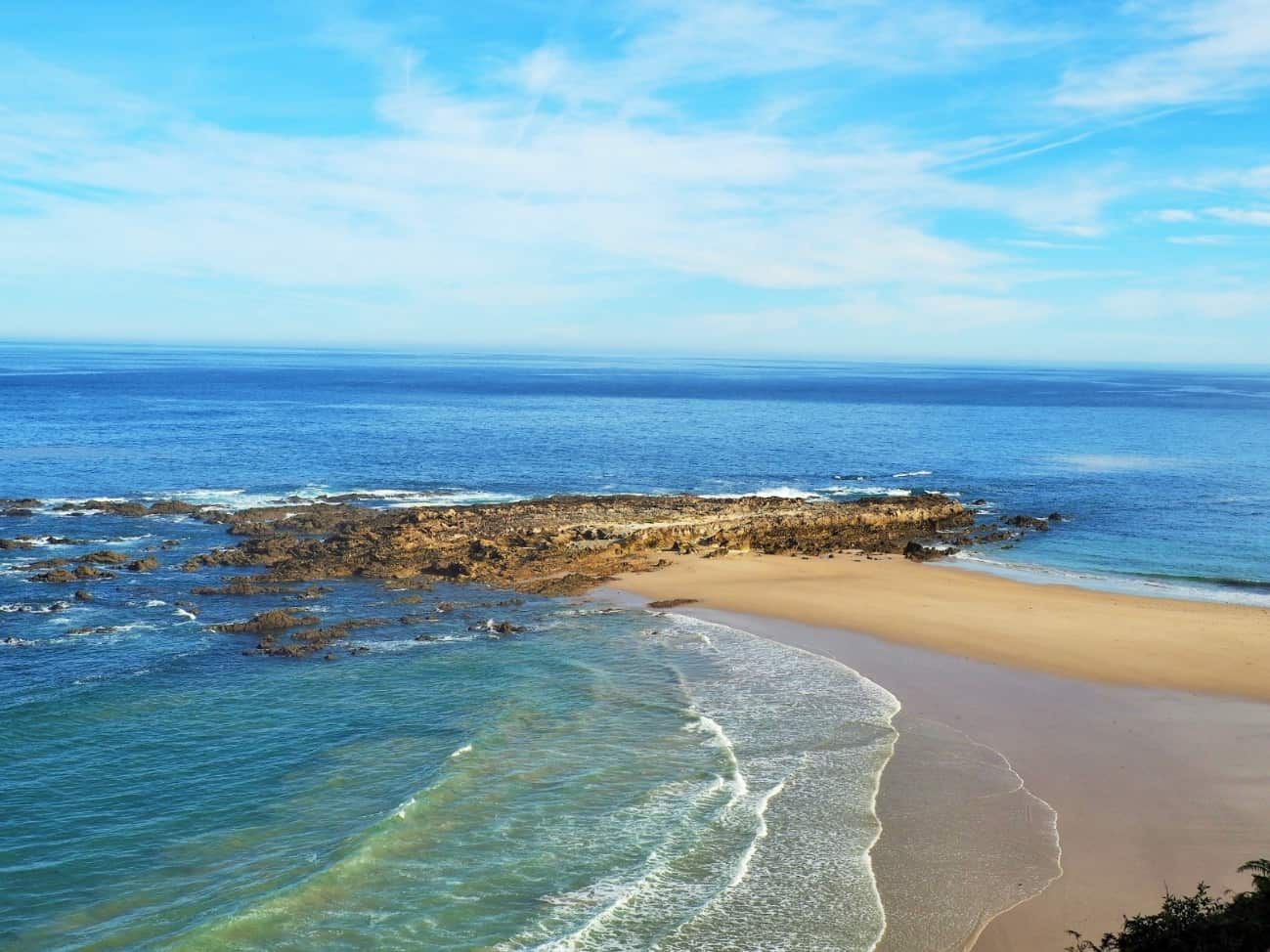 El océano azul y el cielo se encuentran con una playa de arena con afloramientos rocosos.