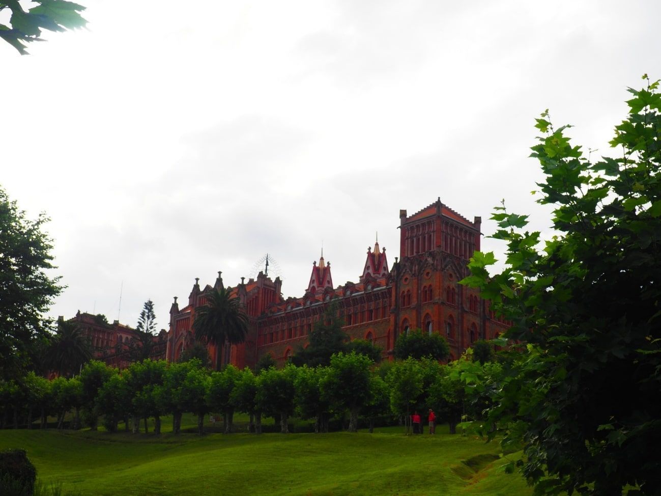 Edificio de ladrillo rojo con torres ornamentadas, visto a través de los árboles en un césped verde bajo un cielo nublado.