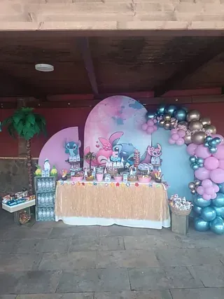 Mesa de postres de cumpleaños con arco de globos rosas y azules y fondo de dibujos animados en un patio cubierto.