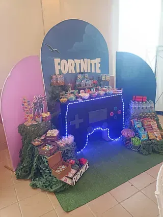 Decoración de fiesta con temática de Fortnite, con un fondo de mando de videojuegos iluminado en azul, mesa de postres y adornos coloridos.