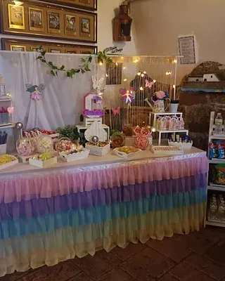 Mesa de postres decorada con falda de arcoíris en tonos pastel, pasteles, flores y luces de hadas en un ambiente festivo en el interior.