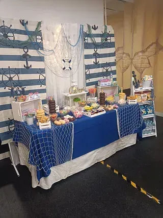 Exhibición de mesa de postres en azul y blanco con bocadillos, bebidas y fondos decorativos con cortinas en un stand de eventos.