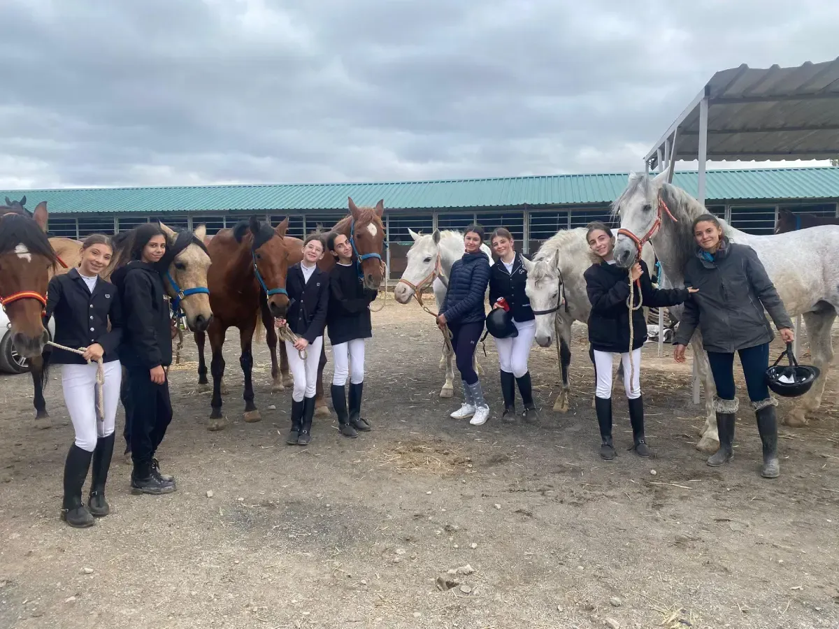 Grupo de jinetes y caballos de pie afuera del establo bajo un cielo nublado.