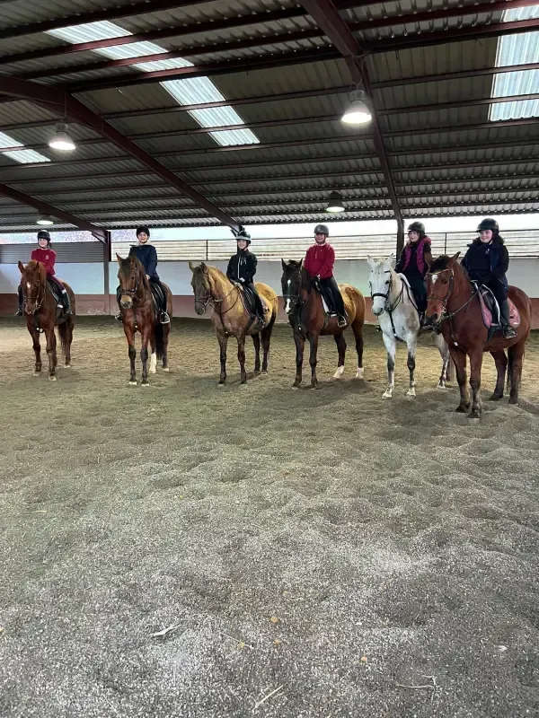 Caballos y jinetes en un picadero cubierto. Varios caballos están de pie, los jinetes sentados.
