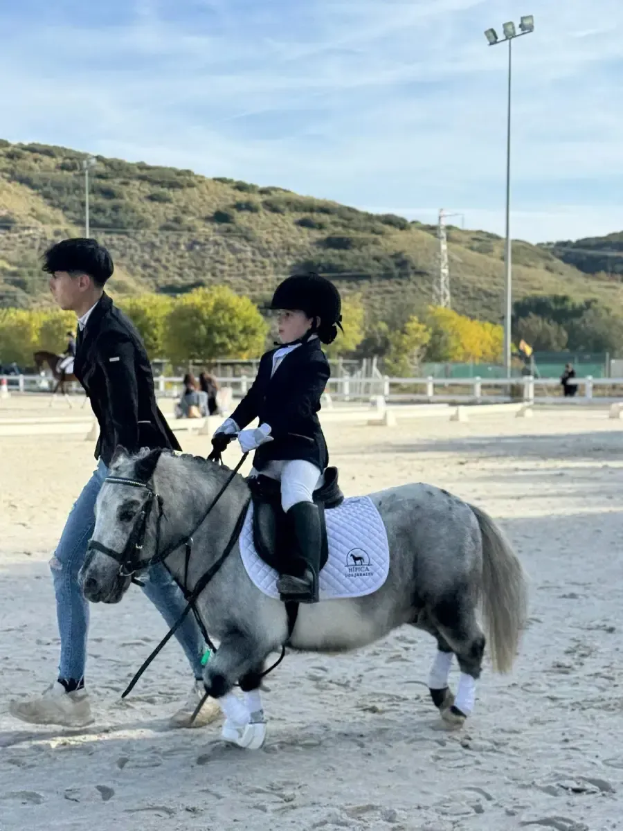 Niño jinete sobre un poni gris, guiado por un guía en una pista. Ambos visten ropa de montar.