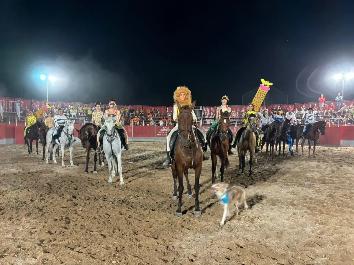Una arena nocturna con jinetes a caballo. Algunos llevan disfraces. Un perro camina delante. El público al fondo.