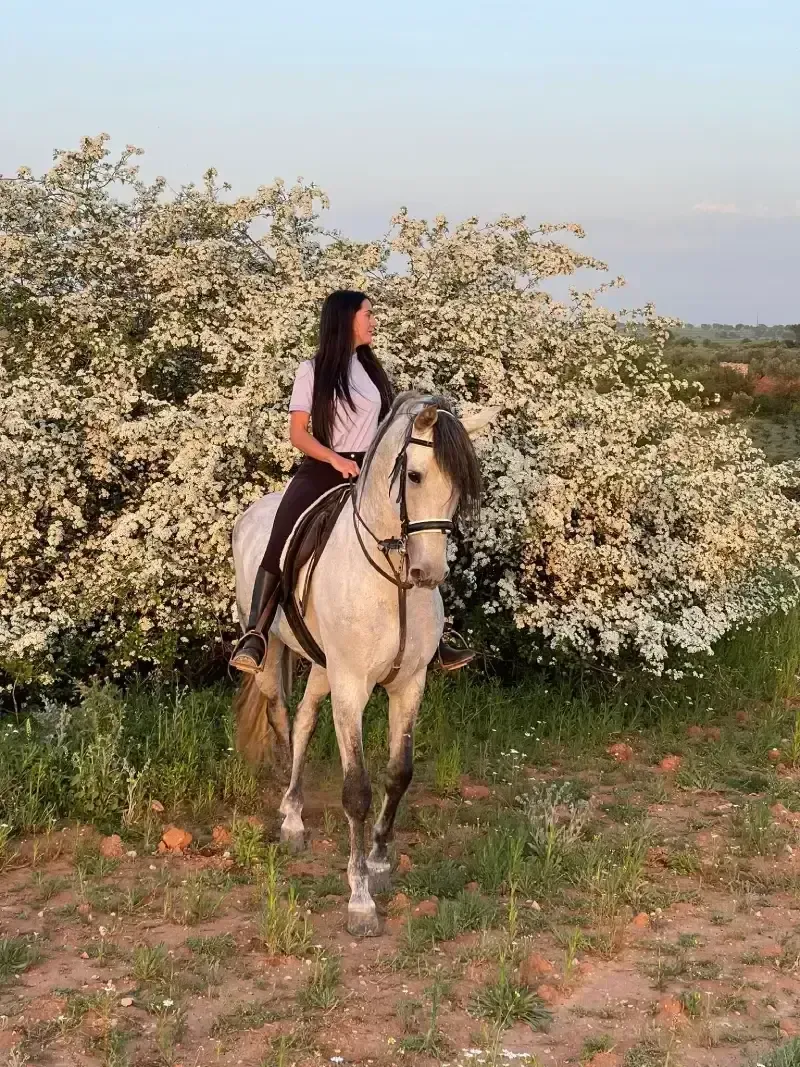 Mujer a caballo frente a flores blancas. El caballo es gris y la mujer tiene el pelo largo y oscuro.