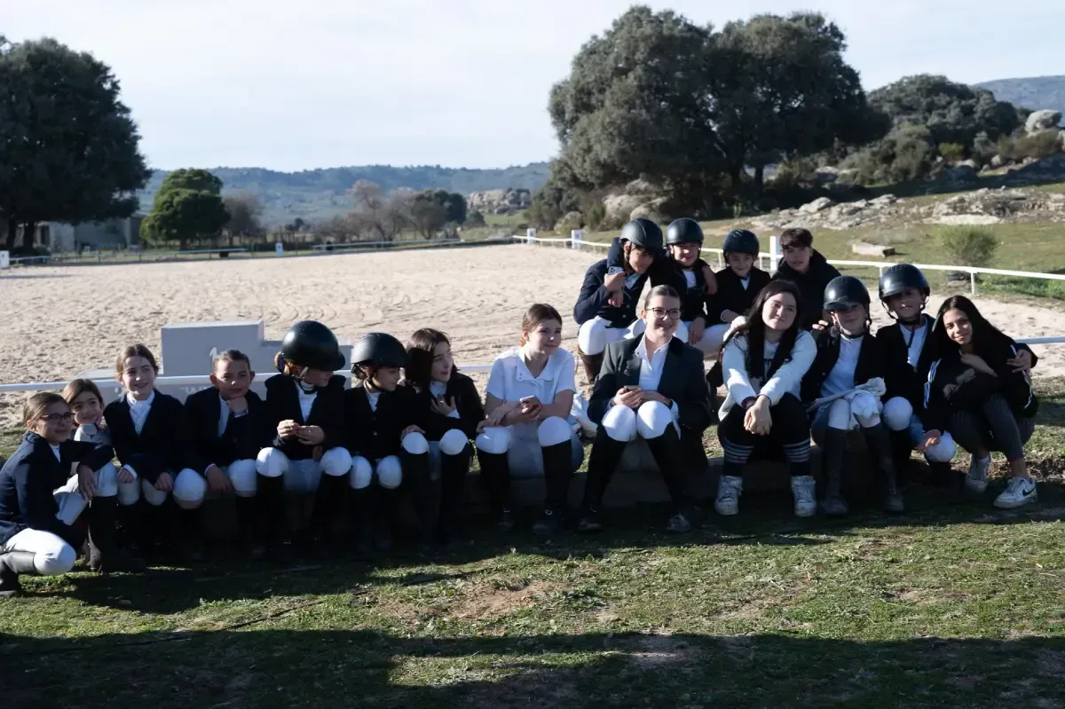 Grupo de jinetes con cascos y ropa de montar, posando sobre el césped con una pista de equitación al fondo.