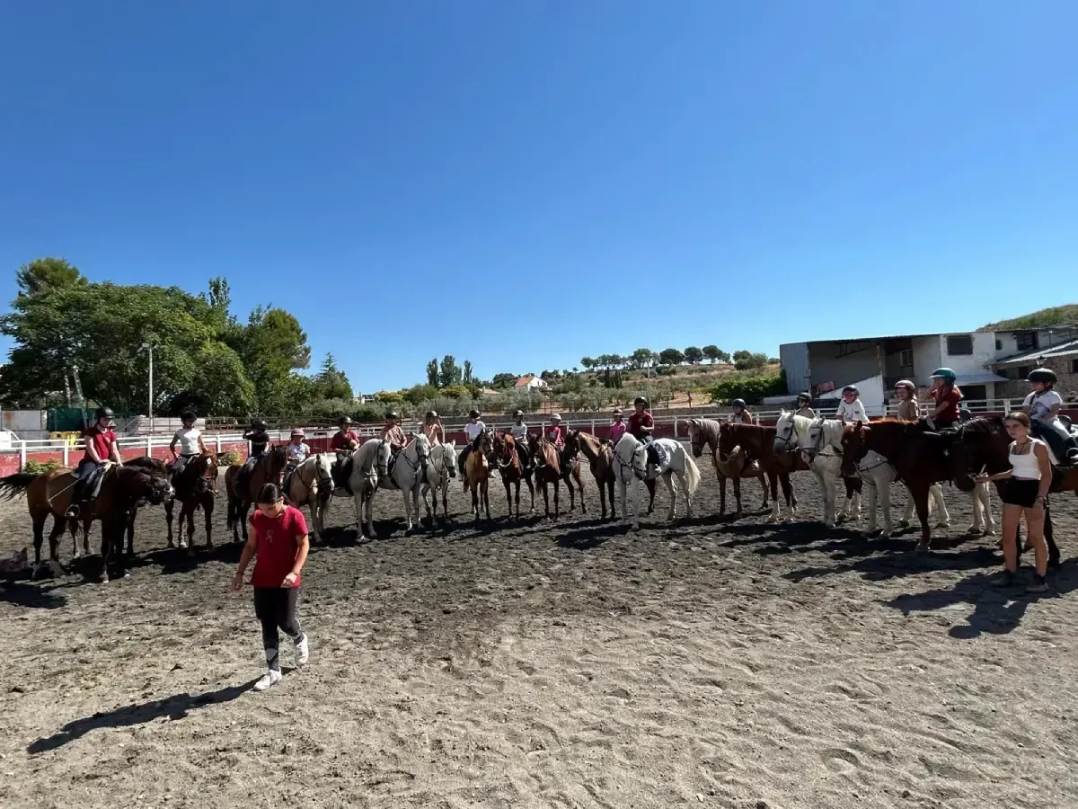 Caballos con jinetes en una arena de tierra bajo un cielo azul, varias personas.