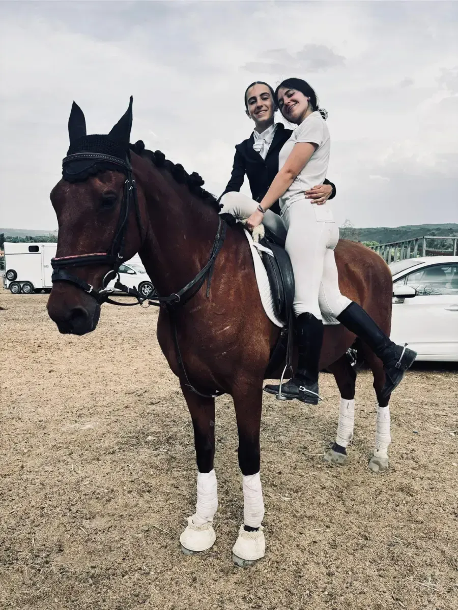 Dos personas en un caballo marrón, una cabalgando, la otra sentada detrás. Escenario ecuestre, cielo nublado.