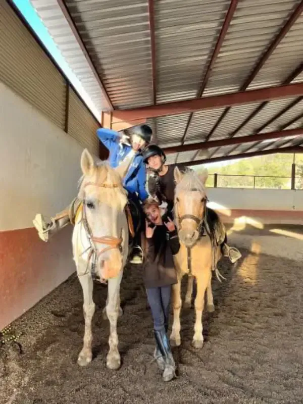 Tres personas a caballo posando, una en cada caballo y otra de pie en el medio, en un picadero cubierto.
