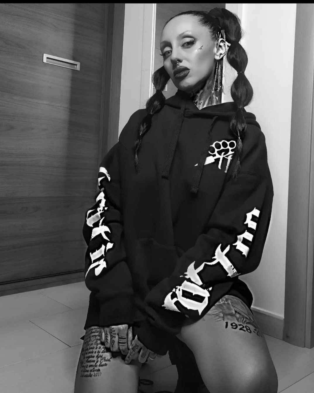 En un retrato en blanco y negro, una persona tatuada con una sudadera negra con mangas estampadas posa arrodillada en el interior de un edificio.