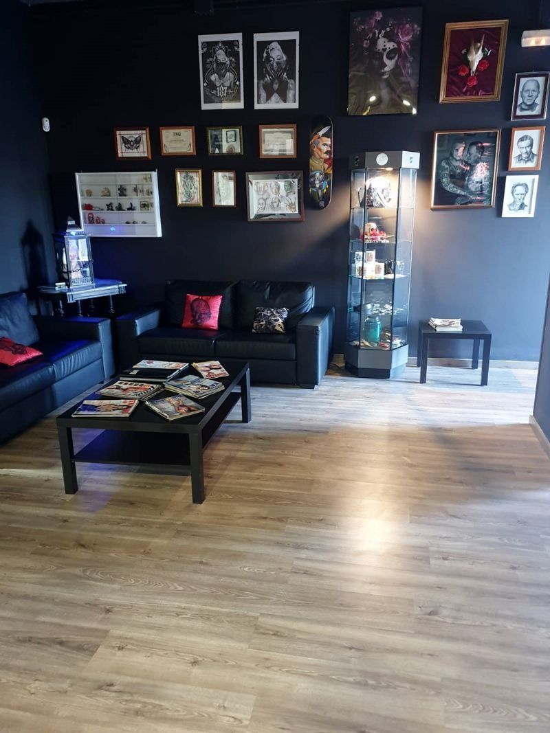 Una sala de espera de un estudio de tatuajes con sofás de cuero negro, una mesa de centro, una vitrina de cristal y cuadros enmarcados en la pared.