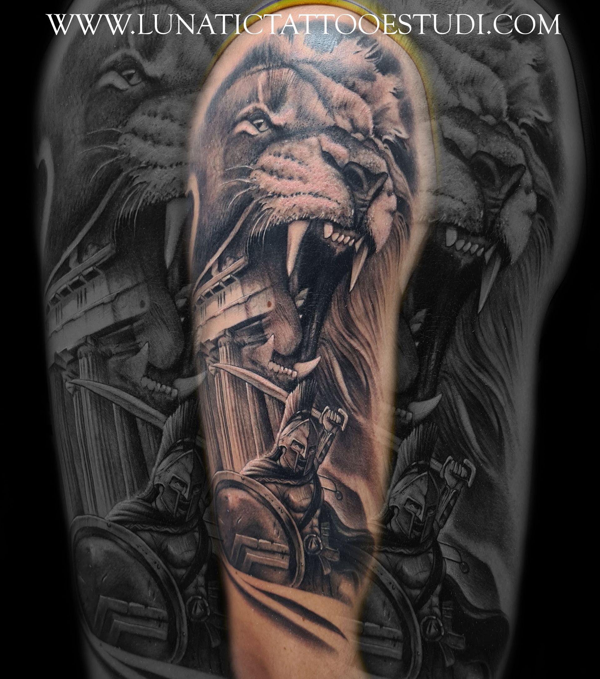 Un tatuaje en blanco y negro en un brazo que muestra un león rugiente que se cierne sobre una escena de batalla con guerreros que portan escudos.