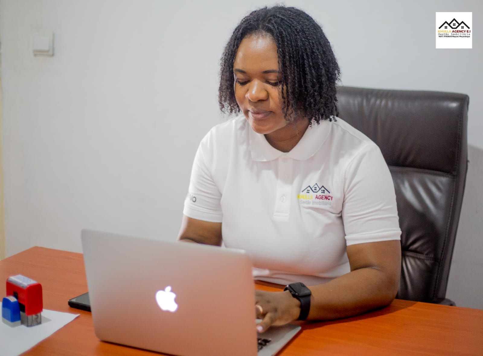 Woman in white polo shirt working on a laptop at a desk, with stamps and a phone nearby.