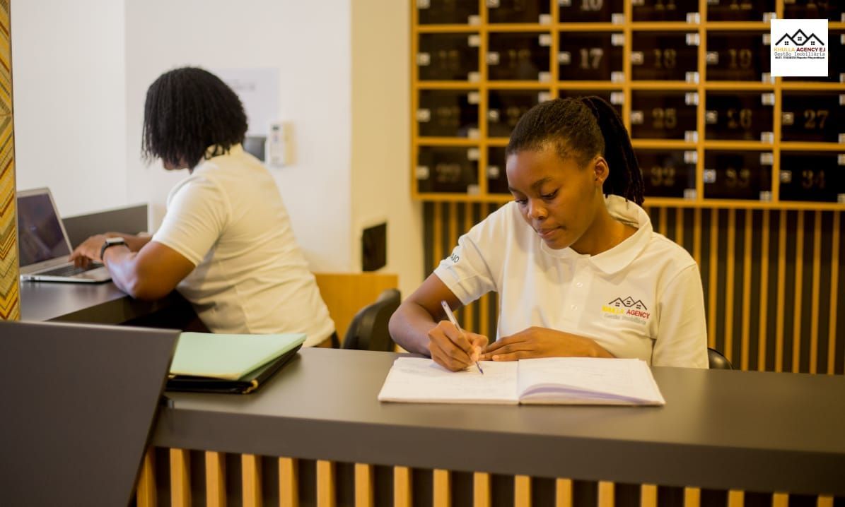 Two women in white shirts at a reception desk; one types on a laptop, the other writes in a notebook.