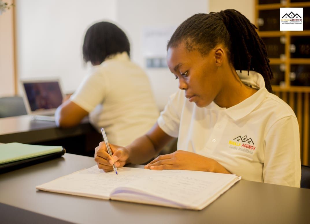 Woman writing in notebook at desk, other person in background. Room setting.