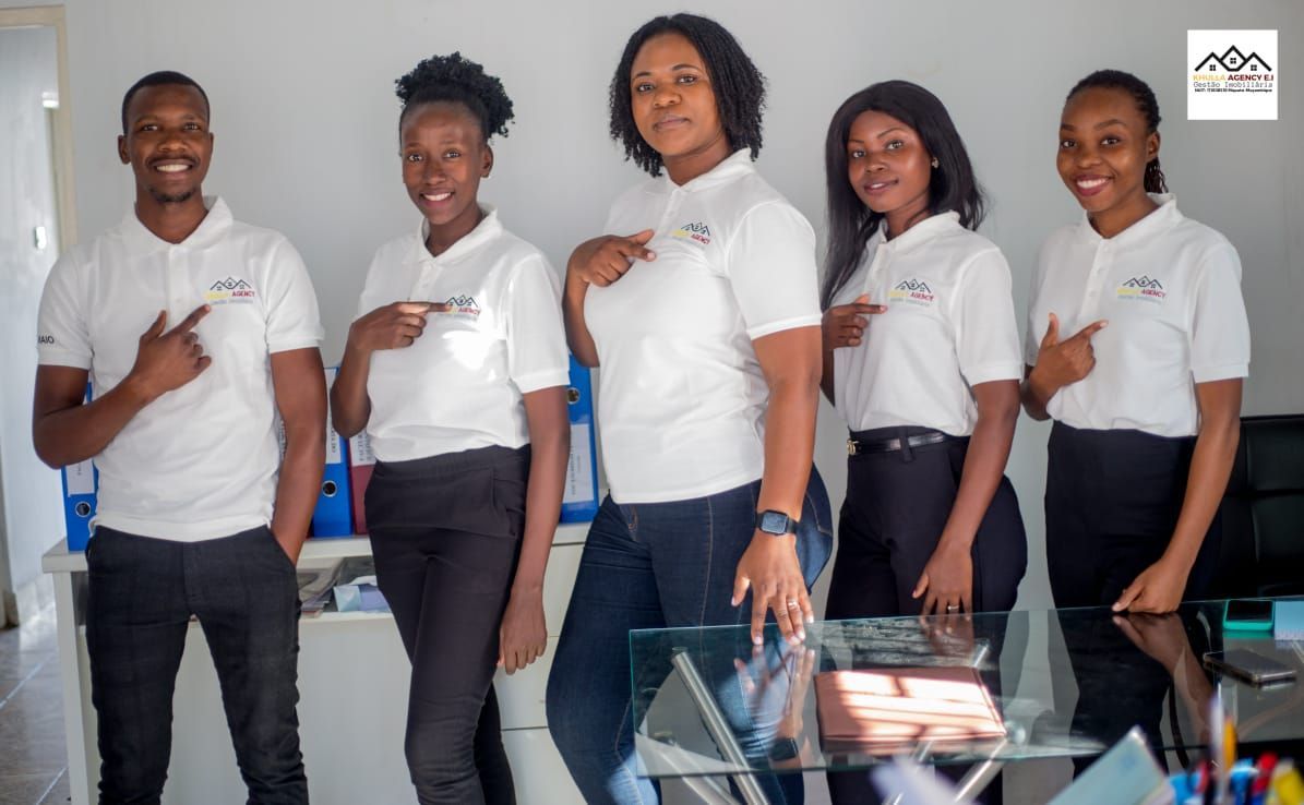 Five people in white shirts and dark pants pointing at themselves, office setting.