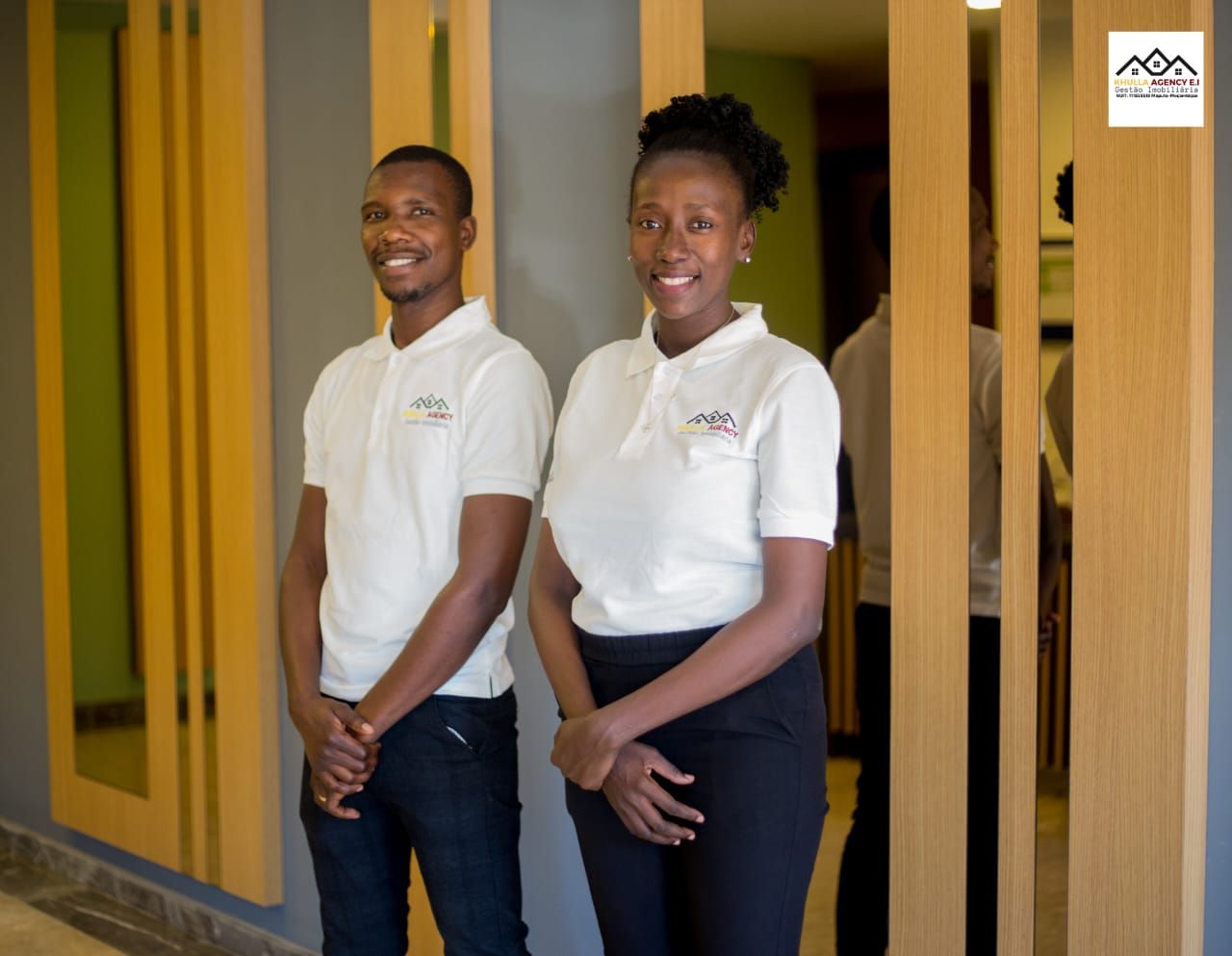 Two people in white polo shirts smile in a hallway with wood and mirror accents.