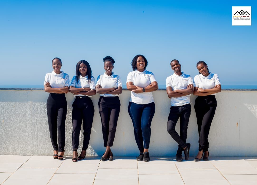 Six women, arms crossed, smiling, wearing white shirts and black pants, posing on a rooftop against blue sky and water.